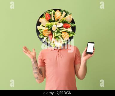Young tattooed man with phone and plate of tasty Caesar salad instead of his head on green background Stock Photo