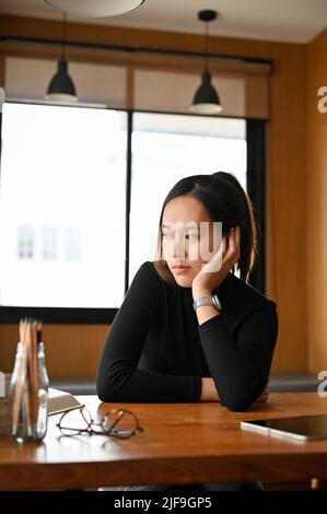 Tired and bored young asian woman or female freelancer looking out the window, pensive thinking something, sitting in the coffee shop Stock Photo