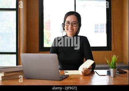 Beautiful young Asian female freelance tutor with eyeglasses and headset sitting in her workspace, preparing to teach her students. Stock Photo