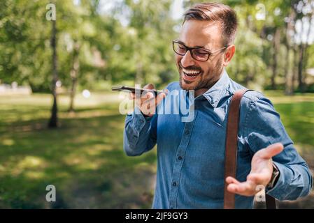 Cheerful male professional talking on smart phone speaker. Young businessman wearing formals is carrying laptop bag. He is walking against trees at pu Stock Photo