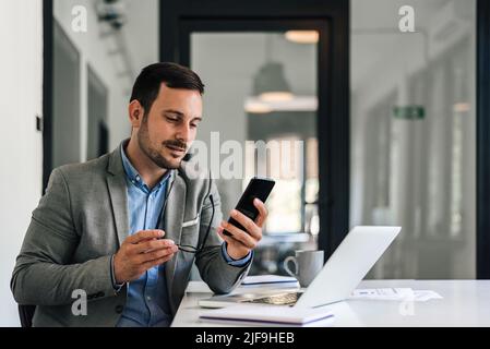 Close-up of confident male executive networking on smart phone. Young entrepreneur with eyeglasses reading messages on cellphone. He is working on lap Stock Photo