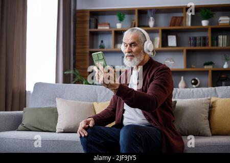 Happy handsome elderly gray-haired man in white headphones holding smartphone and laughing. Old man sitting on couch at home watching funny content Stock Photo