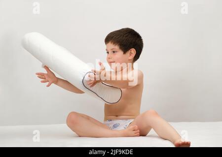A 5-year-old boy tosses an orthopedic pillow of memory foam in the air sitting on a mattress on a white background Stock Photo