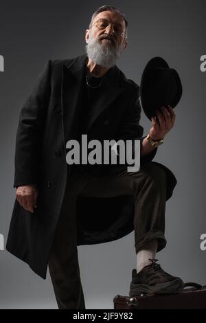 bearded senior man in stylish coat holding derby hat and putting leg on suitcase on grey Stock Photo