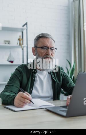 bearded senior man holding pen and using laptop while working from home Stock Photo