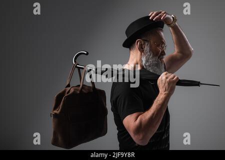 side view of stylish senior man in derby hat holding umbrella with leather bag on grey Stock Photo