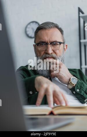 thoughtful bearded senior man in eyeglasses looking at laptop while touching book with hand Stock Photo
