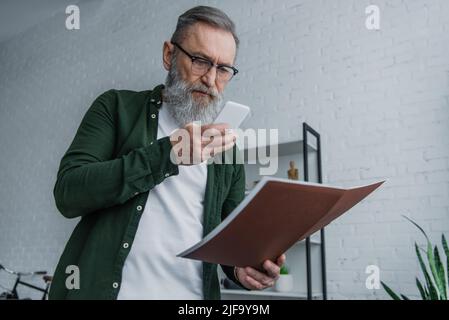 bearded senior man in eyeglasses taking photo of folder on smartphone Stock Photo