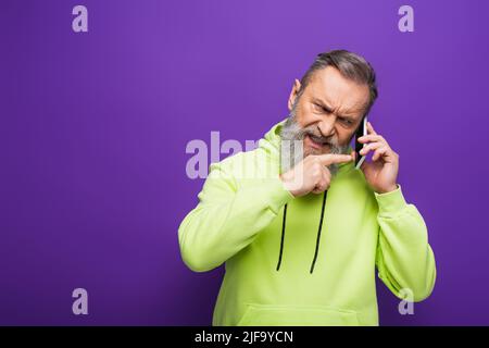 angry senior man in green hoodie pointing with finger while talking on smartphone on purple Stock Photo