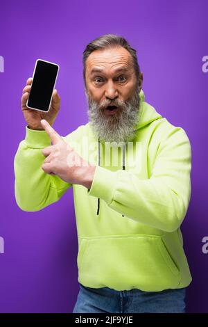 shocked senior man with beard and grey hair pointing at smartphone on purple Stock Photo