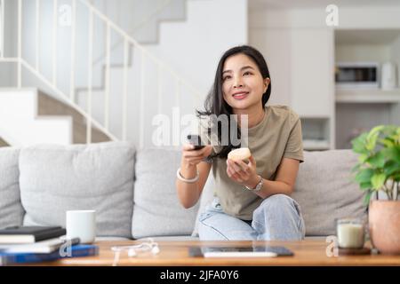 Image of a beautiful asian woman searching channel with remote control to watch tv while sitting on sofa at home Stock Photo