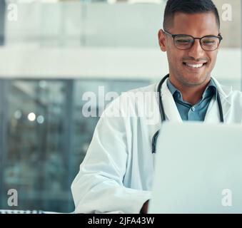 Medical doctor working on his laptop. Young doctor typing on his laptop in the hospital. Smiling doctor browsing the internet on his laptop. Young man Stock Photo