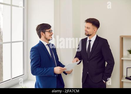Two happy young men in suits standing in office and discussing business contract Stock Photo