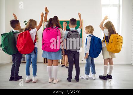Children with large backpacks on their shoulders raise their hands talking with teacher in classroom Stock Photo