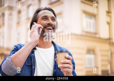 Happy traveling mature bearded man talking using mobile phone and drinking coffee using disposable paper cup standing outdoors in old city background wearing jeans shirt. Freelancer traveling man.  Stock Photo