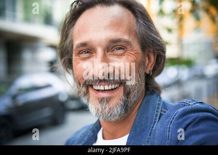 Happy senior man on street of european city making selfie using mobile phone. Middle aged handsome grey bearded man smiling on camera old town wearing casual. Travel concept.  Stock Photo