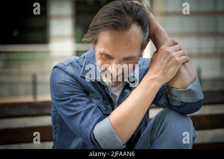Mature grey bearded man deep in his thoughts with positive emotions on his face. Happy thinking of life middle aged man leaned his hands on knee looking down his head sitting on the bench in old town. Stock Photo