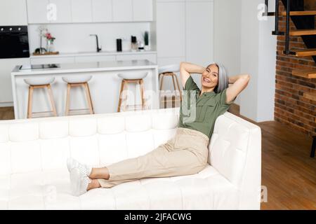 Senior female resting at home. Serene carefree middle-aged woman with long gray hair sitting on sofa in modern apartment, put hands behind head and leaned back Stock Photo