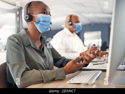 African american call centre telemarketing agent wearing face mask as health and safety protocol talking on headset while using computer in an office Stock Photo