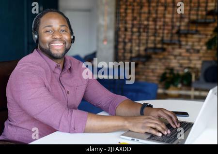 Friendly and positive African-American guy wearing headset using laptop computer working remotely from home, smiling black freelancer man looking at camera sitting in stylish loft apartment Stock Photo