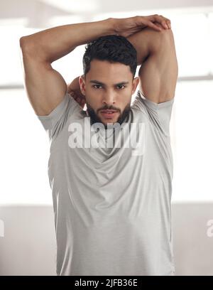 Portrait of one fit young hispanic man stretching arms and shoulders for warmup to prevent injury while exercising in a gym. Serious muscular guy Stock Photo