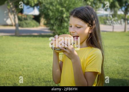 Funny teen girl with a burger in her hands on the background of the park. Stock Photo