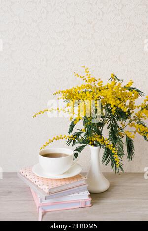 A beautiful composition of mimosa branches and a white tea cup with a saucer and a stack of notebooks on a table with a copy space Stock Photo