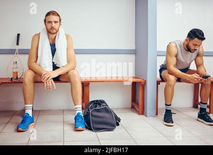 Portrait of a caucasian athlete sitting in a locker room. Two men sitting in a gym locker room together. Serious player relaxing in a gym together Stock Photo