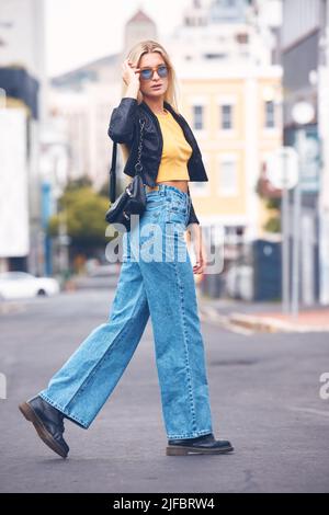 One fashionable young caucasian woman wearing stylish sunglasses while walking in a street in the city. Trendy blonde female looking carefree, cool Stock Photo