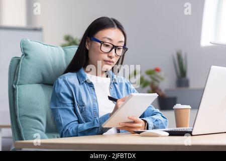 Young Asian woman studying online while sitting in office, writing lecture notes in notebook, using laptop for studying. Stock Photo
