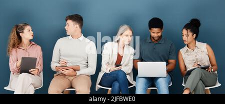 Group of diverse serious businesspeople sitting on chairs in a row in an office together. Five business professionals sitting and talking using Stock Photo