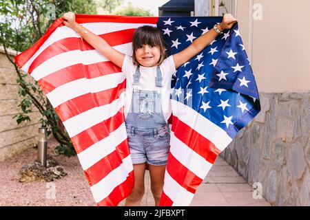 Black-haired girl covered with a large American flag, in the garden of her house. Concept of celebration, independence day, United States of America, Stock Photo