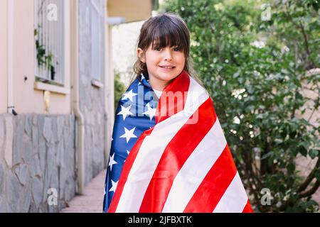 Black-haired girl covered with a large American flag, in the garden of her house. Concept of celebration, independence day, United States of America, Stock Photo