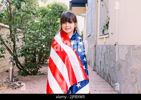 Black-haired girl covered with a large American flag, in the garden of her house. Concept of celebration, independence day, United States of America, Stock Photo