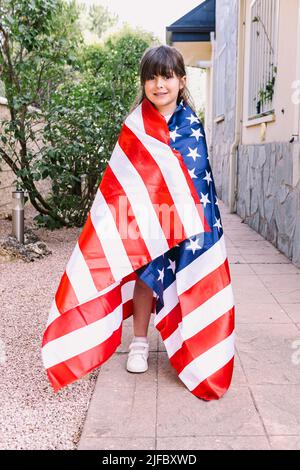 Black-haired girl covered with a large American flag, in the garden of her house. Concept of celebration, independence day, United States of America, Stock Photo