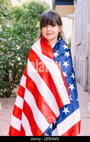 Black-haired girl covered with a large American flag, in the garden of her house. Concept of celebration, independence day, United States of America, Stock Photo