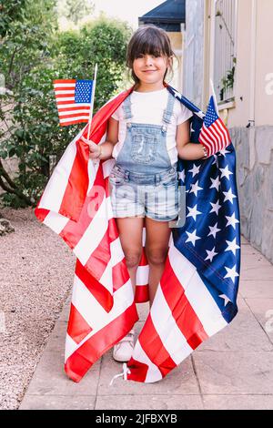 Little black-haired girl covered with big USA flag, holding little American flags in her home garden. Concept of celebration, independence day, 4th of Stock Photo