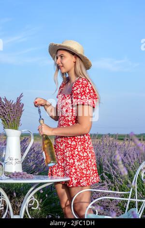 A beautiful young woman opens a bottle of wine in a lavender field on a sunny summer afternoon. Stock Photo