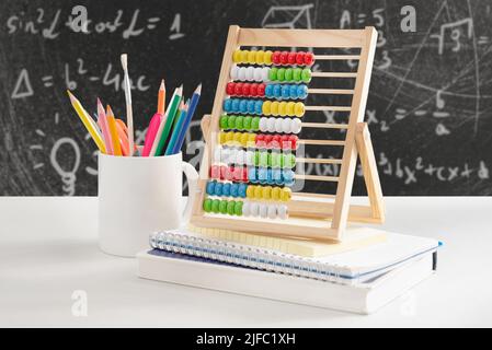 Crayons in mug, abacus and notebooks on school teacher's desk. Blackboard in background Stock Photo