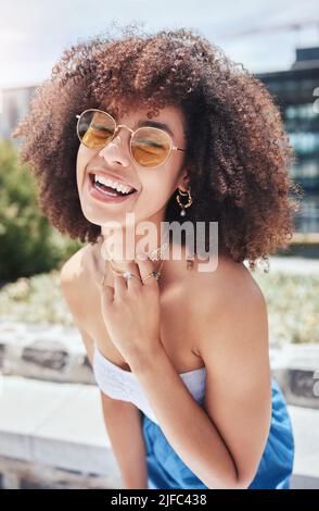 Portrait of young trendy beautiful mixed race woman with an afro smiling and laughing alone outside in the city. Hispanic woman wearing sunglasses and Stock Photo