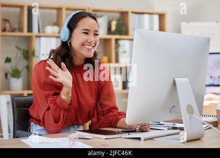 Young happy mixed race woman sitting at her desk at work and using her wireless headphones for an online meeting . Asian businesswoman making a waving Stock Photo