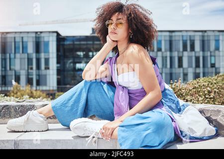 Portrait of a young beautiful stylish african american woman sitting alone outside with yellow sunglasses. Trendy mixed race woman with curly afro Stock Photo