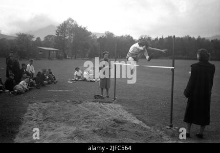 1954, historical, outside in a section of a large field, a girl doing the high jump watched by two lady teachers standing by the horizontal bar or pole set on two posts by a sandpit, Scotland, UK. The young female athlete is using a feet first technique, known as the scissors jump to get over or clear the bar, a style most often by junior athletes and where the landing area was made of sand. Stock Photo