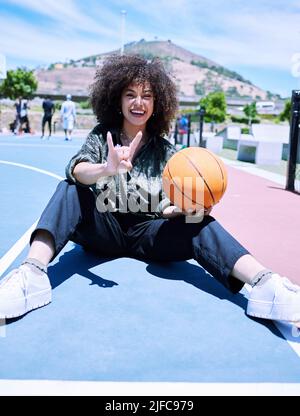 Trendy young hispanic woman making the rock gesture with her hands and fingers, holding a basketball on the court. Happy young woman with afro sitting Stock Photo