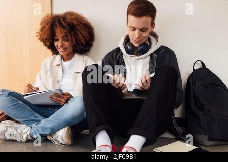 Two classmates sitting at wall in school. Students preparing their assignment while sitting together. Stock Photo