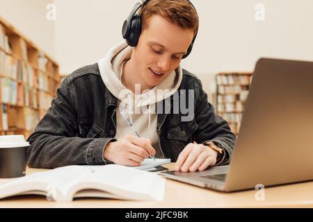 Male student preparing exams at desk in library. Smiling guy in headphones writing on notebook. Stock Photo