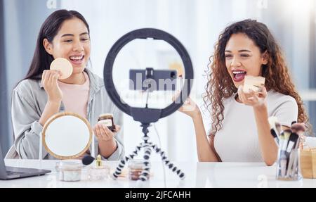 Two mixed race women recording a makeup tutorial for their beauty blog with a phone while sitting at home. Diverse young influencers live streaming Stock Photo