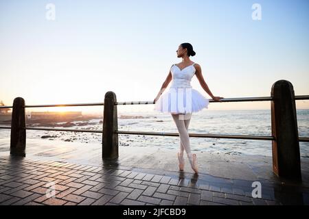 Elegant young ballerina wearing white fancy dress and pumps while leaning against a railing on the promenade with a beautiful ocean background at Stock Photo