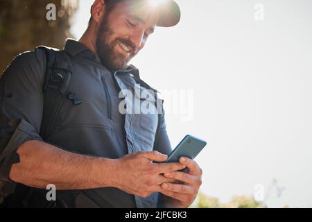 Happy caucasian man using smartphone while carrying a backpack and hiking alone in the mountains on a sunny day. Smiling fit and active man using Stock Photo