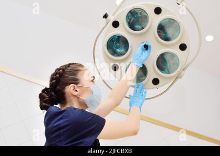 attractive female medic in a sterile mask and rubber gloves turns on a surgical lamp in a modern operating room. Stock Photo